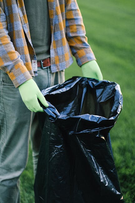 A pile of mixed waste materials, including flattened cardboard boxes, some with printed logos and barcodes, and plastic packaging, situated on the ground near a brick wall and a rough stone base. The
