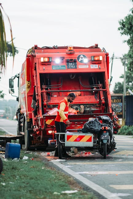 A sanitation worker is operating a large red rubbish collection vehicle on a street near a curb, with its rear hatch open, revealing mechanical components and storage space for waste. The worker, dres